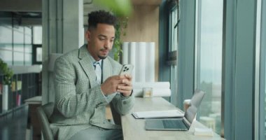 Ekose takım elbiseli genç bir işadamı akıllı telefonunu kullanarak modern bir ofis masasında oturuyor. Bir dizüstü bilgisayar, kahve fincanı ve belgeler masanın üzerinde, geniş pencereli, parlak, profesyonel bir ortam sağlıyor.