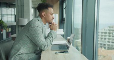 Kareli ceketli profesyonel bir adam masanın üzerine eğilir ve laptopunun yanındaki pencereden dışarı dikkatlice bakar. Görüntü, modern ofis ortamındaki konsantrasyon ve içgözlem yansımalarını yansıtıyor. Yüksek