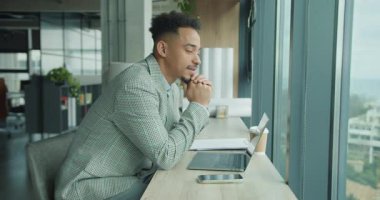 Kareli ceketli profesyonel bir adam masanın üzerine eğilir ve laptopunun yanındaki pencereden dışarı dikkatlice bakar. Görüntü, modern ofis ortamındaki konsantrasyon ve içgözlem yansımalarını yansıtıyor. Yüksek