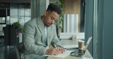 Ekose ceketli profesyonel bir işadamı, modern bir ofis masasındaki bir deftere notlar yazarak derinlemesine odaklanır. Çalışma alanında dizüstü bilgisayar, kahve fincanı ve belgeler var. Yüksek kalite 4k