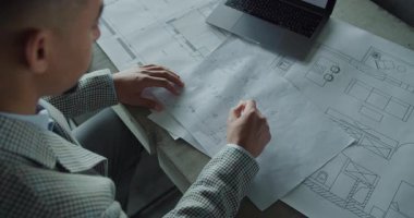 Bir mimar dikkatle inceler ve yakınındaki bir dizüstü bilgisayarla beton bir masanın planlarını ve çizimlerini işaretler. Görüntü kesinlik, planlama ve tasarım odaklı yaratıcılığı vurguluyor. Yüksek kalite 4k