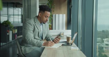 Ekose ceketli bir işadamı modern bir ofis masasında çalışıyor. Bir dizüstü bilgisayar, kahve ve belgelerle çevrili akıllı telefonunu kontrol ederken bir deftere yazıyor. Yüksek kalite 4k görüntü