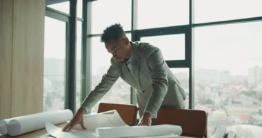 Bir işadamı, şehir manzarasına bakan geniş pencereli modern, aydınlık bir ofisin planlarını yakından analiz ederek masanın üzerine eğiliyor. Çalışma alanı gösterişli ve profesyonel. Yüksek kalite