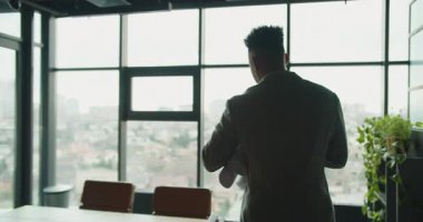 Şehir manzarası olan büyük bir ofis penceresinin yakınındaki planları inceleyen profesyonel bir işadamı. Modern ofis doğal ışık ve endüstriyel tasarıma sahiptir. Yüksek kalite 4k görüntü