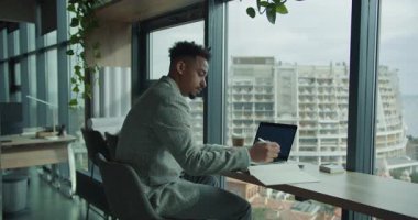 Ekose ceketli profesyonel bir işadamı modern bir ofis masasında çalışır ve bilgisayarındaki verileri analiz eder. Ofiste şehir manzaralı büyük pencereler var, profesyonel bir atmosfer yaratıyor.