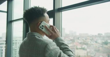Modern bir ofisin geniş pencerelerinden dışarı bakarken şık, damalı ceketli bir iş adamı telefon görüşmesi yapıyor. Şehir manzarası profesyonel bir zemin sağlar. Yüksek kalite 4k görüntü
