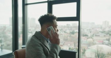 Profesyonel bir işadamı, şehir manzarasına bakan geniş pencereli modern bir ofiste telefon görüşmesi yapıyor. Adam şık, kareli bir ceket giyiyor. Yüksek kalite 4k görüntü