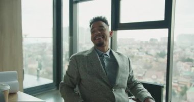 Dama desenli ceketli neşeli bir işadamı sandalyesine yaslanmış, şehir manzarasına bakan geniş pencereli, parlak ve modern bir ofise güvenle gülümsüyor. Yüksek kalite 4k görüntü