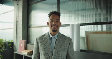 Kareli bir takım elbise giyen kendine güvenen bir işadamı, arka planda kitaplar, bitkiler ve mimari planlarla modern bir ofis ortamında dururken gülümsüyor. Yüksek kalite 4k görüntü