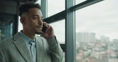 Ekose takım elbiseli kendine güvenen bir işadamı modern bir ofis penceresinin yanında duruyor, telefonda konuşuyor, arka planda bulanık bir şehir manzarası var. Yüksek kalite 4k görüntü