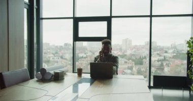 Modern bir ofis masasında dizüstü bilgisayarı, kahve fincanı ve planları olan profesyonel bir işadamı telefonda konuşuyor. Büyük pencereler şehir manzarası manzarası sağlar. Yüksek kalite 4k görüntü
