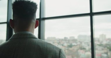 Modern bir ofisin geniş pencerelerinin yanında duran ekose takım elbiseli profesyonel bir işadamının uzaktan kentsel manzarayı gözlemlemesi. Yüksek kalite 4k görüntü