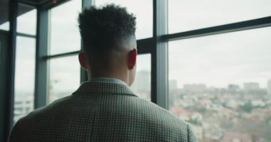 Modern bir ofis penceresinin yanında kareli takım elbiseli profesyonel bir işadamının uzaktan bulanık şehir manzarasını gözlemlemesinin yan görüntüsü. Yüksek kalite 4k görüntü