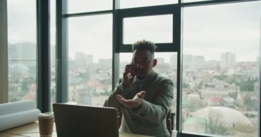 Modern bir ofisteki profesyonel bir işadamı telefonda konuşarak ve dizüstü bilgisayar kullanarak birden fazla iş yapıyor. Arka planda şehir manzarası olan bir kahve fincanı ve planlar masanın üzerinde duruyor. Yüksek
