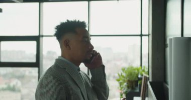 Ekose takım elbiseli profesyonel bir işadamı modern bir ofiste telefonla konuşuyor. Çalışma alanında şehir manzaralı büyük pencereler, yeşil santraller ve mimari planlar yer alıyor. Yüksek kalite 4k