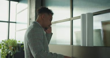 Ekose takım elbiseli profesyonel bir işadamı modern bir ofiste masanın yanında dururken telefonla konuşuyor. Çalışma alanını mimari planlar ve yeşil bitkiler dekore ediyor. Yüksek kalite 4k