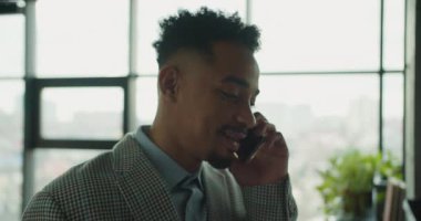 Parlak bir ofiste telefonla konuşurken gülümseyen ekose takım elbiseli profesyonel bir iş adamı. Büyük pencereler ve yeşillikler misafirperver ve şık bir ortam yaratır. Yüksek kalite 4k görüntü