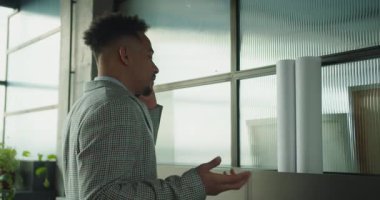 Ekose takım elbiseli profesyonel bir işadamının modern bir ofisteki telefon görüşmesinde el kol hareketi yapması. Arka planda mimari planlar ve desenli cam paneller var. Yüksek kalite 4k