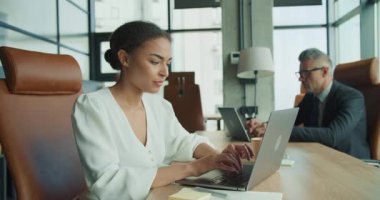 Ortak bir ofis masasında dizüstü bilgisayarda çalışan kendine güvenen bir iş kadını. Arka planda takım elbiseli bir işadamı çalışıyor. Doğal gün ışığı ve odaklanma ile modern çalışma alanı.