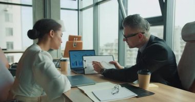 İşadamı, modern yüksek katlı bir ofiste dizüstü bilgisayarlı ve kahveli odaklı bir toplantı sırasında, pano kullanan bir bayan meslektaşına mali hat çizelgesini açıklıyor..