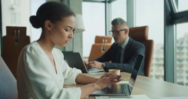 Genç iş kadını dizüstü bilgisayarla çalışırken, iş adamı arka planda bir şeyler yazıyor. Geniş pencereleri ve doğal ışığı olan modern ofis parlak bir atmosfer yaratıyor..