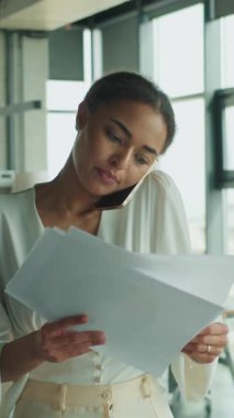 Bir iş kadını parlak bir ofiste duruyor, belgelerini okurken telefonla konuşuyor. Arka planda bir iş adamı çalışıyor. Çoklu görev ve üretkenlik kavramı.