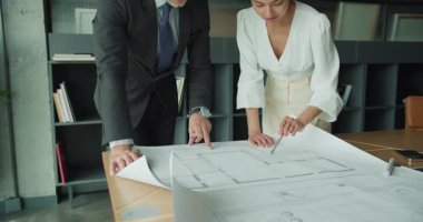 Profesyonel işadamı ve iş kadını modern ofis ortamında bir masada mimari planları tartışıyor, proje planlama ve tasarım stratejisine odaklanıyor.