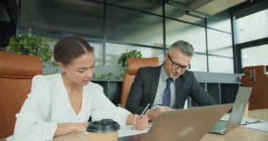 Profesyonel kıyafetli bir erkek ve kadın, fabrikalarla dolu modern bir ofiste iş belgeleri ve dizüstü bilgisayarlar üzerinde yoğun bir şekilde çalışıyorlar. Veri analiz görevlerine odaklanmışlardır..