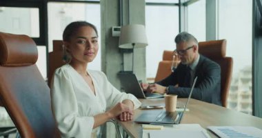 Kendine güvenen iş kadını, erkek arkadaşı arkasında çalışırken modern bir ofiste masasında poz veriyor. Parlak gün ışığı, dizüstü bilgisayarlar, çizelgeler ve kahve profesyonel bir ortam yaratır..