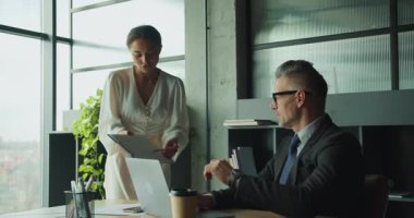 Kendine güvenen bir iş kadını modern bir ofiste dizüstü bilgisayarda çalışan bir iş adamına belgeler gösterir. Takım çalışması, iletişim ve kurumsal karar verme kavramı.