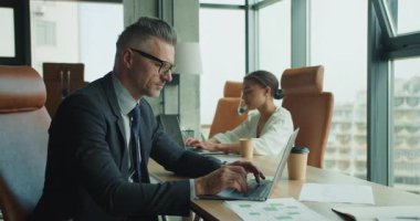 İki iş adamı parlak, modern bir ofiste dizüstü bilgisayarlara odaklandı. Takım elbiseli bir adam ve beyaz bluzlu bir kadın masada evraklar ve kahve fincanlarıyla yan yana çalışıyorlar..