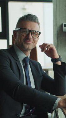 Takım elbiseli kendine güvenen işadamı modern bir ofiste dizüstü bilgisayarla çalışırken kameraya gülümsüyor. Arka plandaki iş kadını kendi görevlerine odaklanmış. Parlak çalışma alanı.