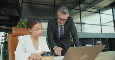 Takım elbiseli iş adamı, modern bir ofisteki kadın meslektaşına takım stratejisi ve veri analiziyle ilgili bir şeyi açıklarken laptop ekranını işaret ediyor..