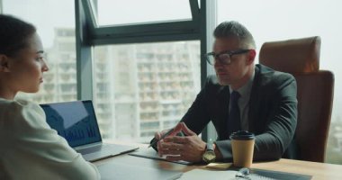 Olgun iş adamı, proje detaylarını meslektaşıyla dikkatlice tartışıyor. Bir dizüstü bilgisayardaki bilgi grafikleri profesyonel ofis ortamını güçlendirir.