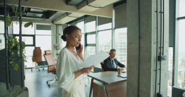 Modern bir ofiste çalışan bir iş kadını, arka planda bir işadamı çalışırken telefonla konuşur ve bir belge okur. Parlak profesyonel çalışma alanı.