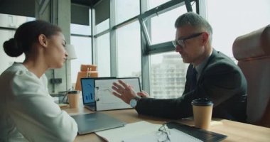 İşadamı, bir iş kadınıyla satış verilerini tartışırken panonun üzerindeki bir çizelgeye işaret ediyor. Dizüstü bilgisayarı ve masasında kahve olan modern ofis..