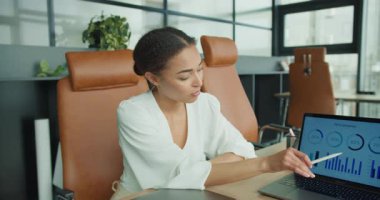 Kendine güvenen iş kadını, toplantı sırasında bilgi tablolarını açıklarken, modern, parlak bir ofis ortamında dizüstü bilgisayardaki verilere işaret ediyor..