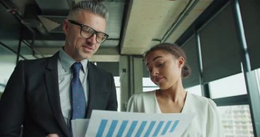 Bir işadamı ve bir iş kadını modern bir ofiste yazılı mali verileri tartışıyor. Profesyonel bir sohbete odaklanmış ve meşgul görünüyorlar..
