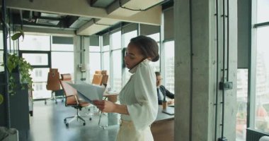 Bir iş kadını parlak bir ofiste duruyor, belgelerini okurken telefonla konuşuyor. Arka planda bir iş adamı çalışıyor. Çoklu görev ve üretkenlik kavramı.