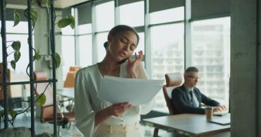 Bir iş kadını, modern bir ofisteki belgeleri incelerken telefonu omzuyla tutarak birden fazla iş yapar. Arka planda bir işadamı elinde kahve fincanıyla oturuyor..