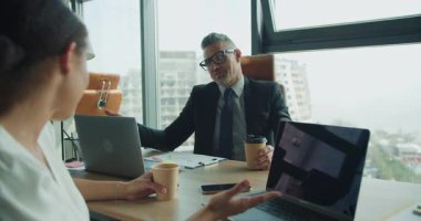 İşadamı ve iş kadını yüksek katlı bir ofiste kahve yakıtlı bir masada sohbet ediyorlar. Dizüstü bilgisayarlar, belgeler ve doğal ışık modern bir çalışma alanı hissi yaratır..