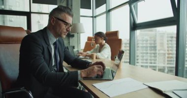 Odaklanmış işadamı ve iş kadını, şehir manzaralı, üretkenliği, takım çalışmasını ve profesyonel çalışma ortamını vurgulayan modern bir ofiste dizüstü bilgisayarlar üzerinde çalışıyorlar..