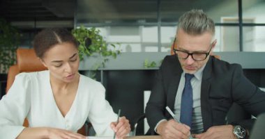 İş adamı ve iş kadını, modern bir ofisteki basılı çizelgeleri analiz etmek için masa başında yakın çalışırlar. Odaklanmış ifadeler ayrıntılı ve işbirlikçi bir çalışma oturumu önerir.