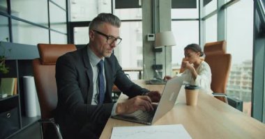 İş kadını modern ofiste büyük pencereli kahve içerken, iş adamı laptopta çalışırken, üretkenlik ve gevşetilmiş işbirliği dengesini gösteriyor.