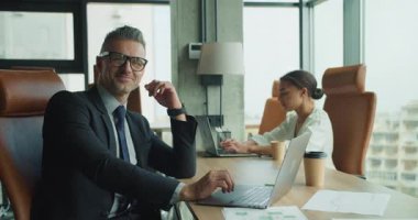 Takım elbiseli kendine güvenen işadamı modern bir ofiste dizüstü bilgisayarla çalışırken kameraya gülümsüyor. Arka plandaki iş kadını kendi görevlerine odaklanmış. Parlak çalışma alanı.