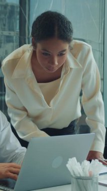 İş kadını, toplantı sırasında dizüstü bilgisayarda çalışan bir meslektaşına yardım etmek için eğiliyor. İkisi de geniş pencereli modern bir ofise odaklanmış ve meşgul..