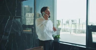 Beyaz gömlekli bir iş adamı büyük bir ofis penceresinin yanında dururken akıllı bir telefonla konuşur. Doğal ışık modern çalışma alanını mobilya manzarasıyla doldurur. Yüksek kalite 4k görüntü