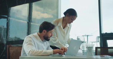 İş adamı dizüstü bilgisayarda yazı yazarken, meslektaşı da büyük camları ve yansımaları olan parlak ve modern bir ofiste yaptığı toplantıda yakından gözlemliyor ve yardımcı oluyor. Yüksek kalite 4k görüntü