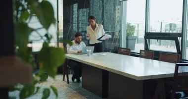 İş adamı dizüstü bilgisayarın başında otururken yanında bir iş kadını elinde belgelerle duruyor. Geniş, güneşli bir ofiste geniş bir konferans masasında birlikte çalışıyorlar. Yüksek kalite 4k görüntü