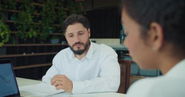 Beyaz gömlekli bir iş adamı toplantı sırasında bir meslektaşını dikkatle dinler. Modern ofis ortamında masada grafikli kağıtlar ve dizüstü bilgisayarlar görülebilir. Yüksek kalite 4k görüntü